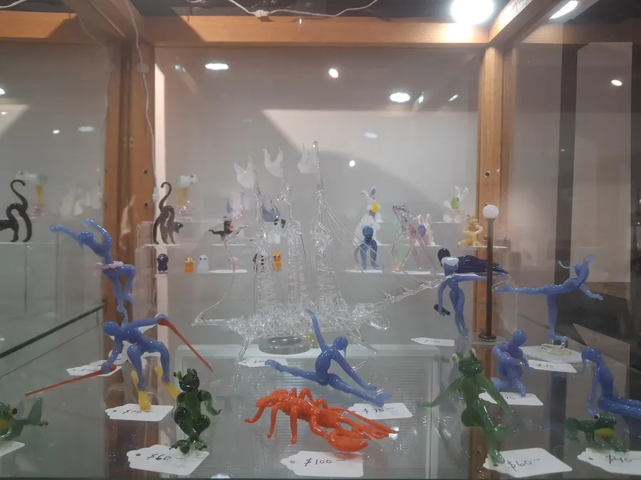 Display case of Elton Clarke glass sculptures including figures, a clear sailing ship, frogs, and a lobster