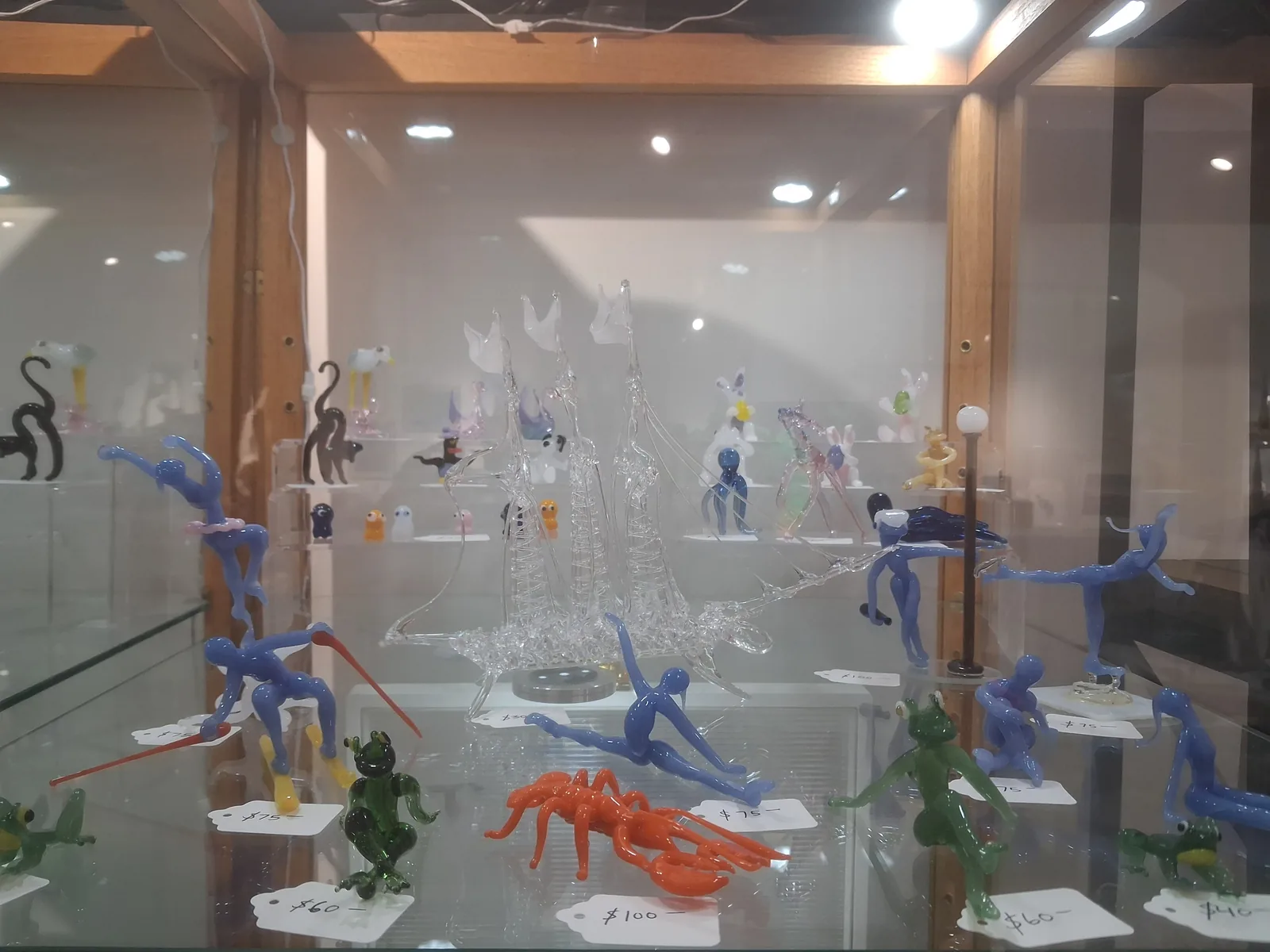 Display case of Elton Clarke glass sculptures including figures, a clear sailing ship, frogs, and a lobster