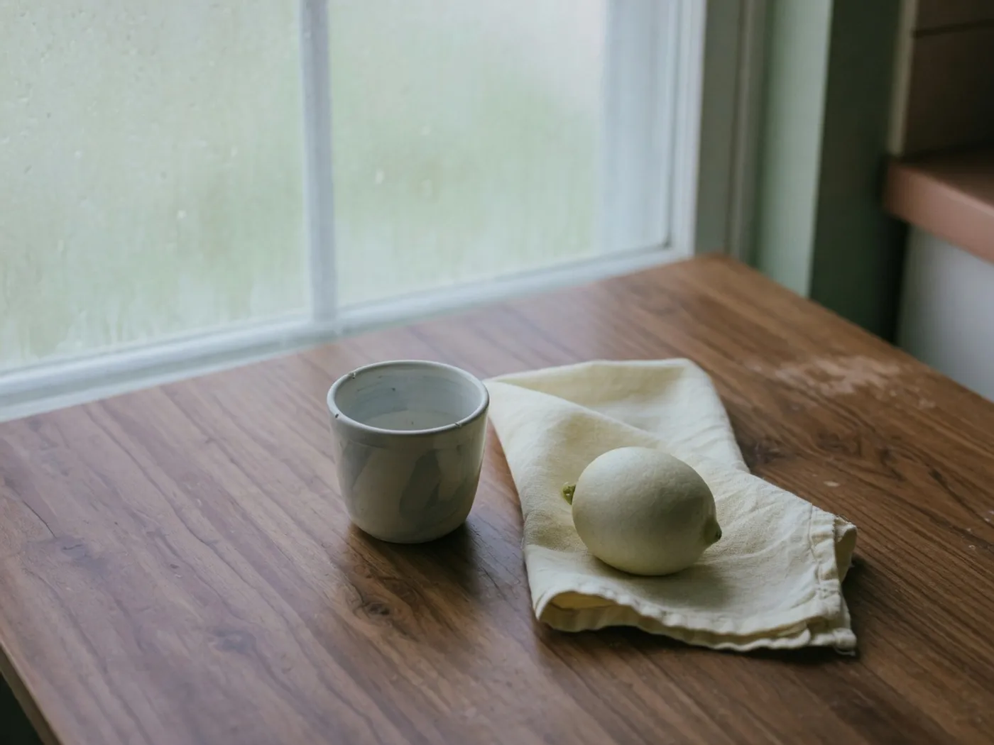A ceramic cup, folded linen, and pale lemon on a walnut kitchen table